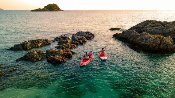 Two people paddle red kayaks through rocky outcrops amid a turquoise sea at sunset